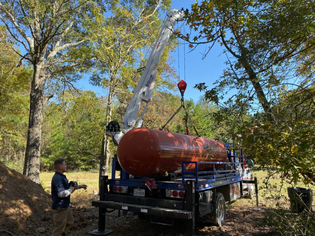 Orange propane tank being handled by a truck-mounted crane beside trees