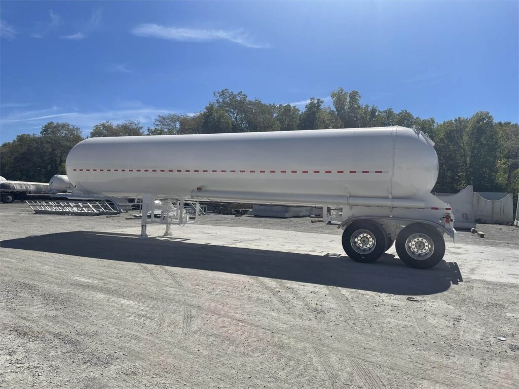 White LPG tank trailer seen from the side, parked on a gravel lot with trees behind